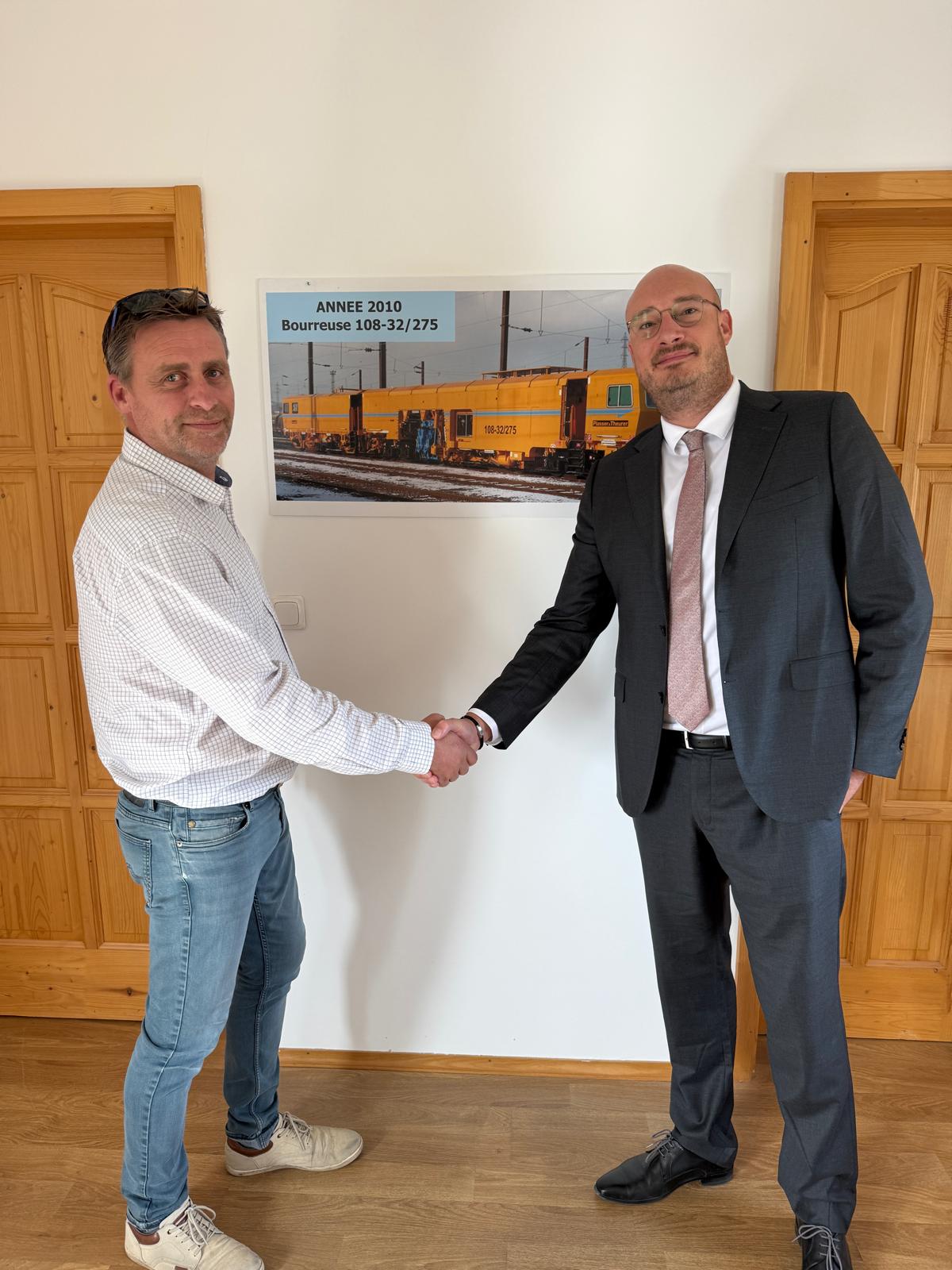 Poignée de main entre deux dirigeants RAILMAT devant une affiche de la bourreuse 108-32/275